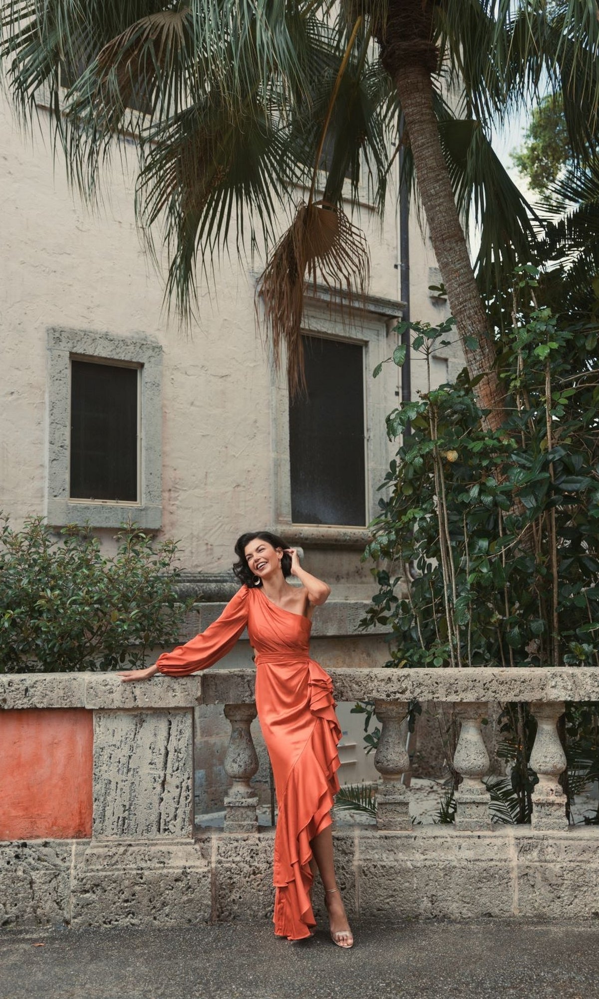 A woman wearing the Long Formal Dress 49531 by Mac Duggal, an orange one-shoulder gown, smiles and touches her hair outdoors by a stone balustrade, with greenery and a stucco building in the background.